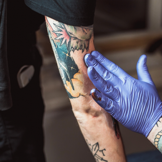 Close-up of an artist wearing purple nitrile gloves applying aftercare cream onto a client's freshly inked colorful arm tattoo.