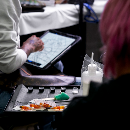 A blurred view of a tattoo artist working on a digital design with a tray of ink and needles in the foreground.