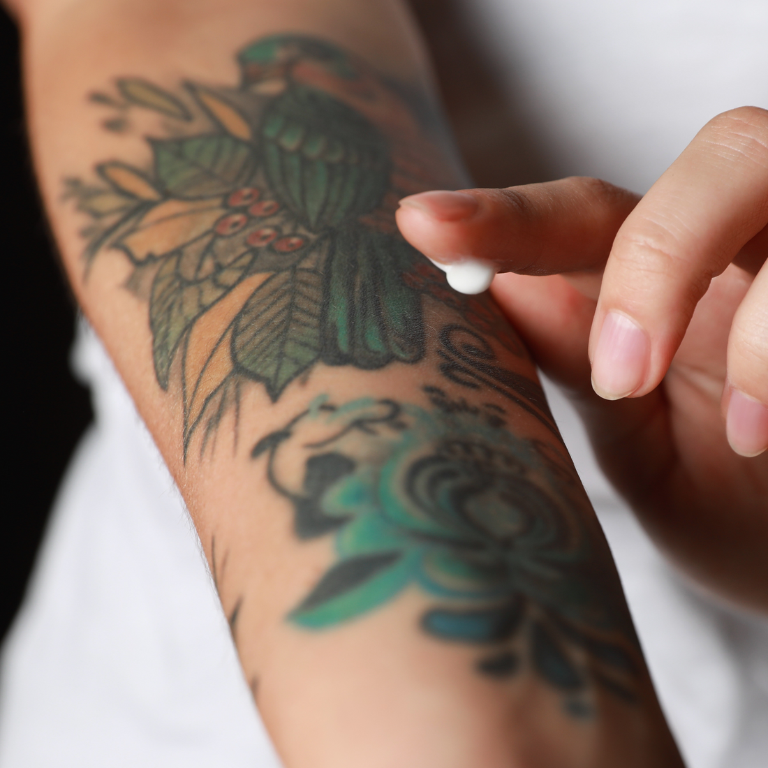 A finger applying a small amount of white soothing lotion to a vibrant bird and floral forearm tattoo.