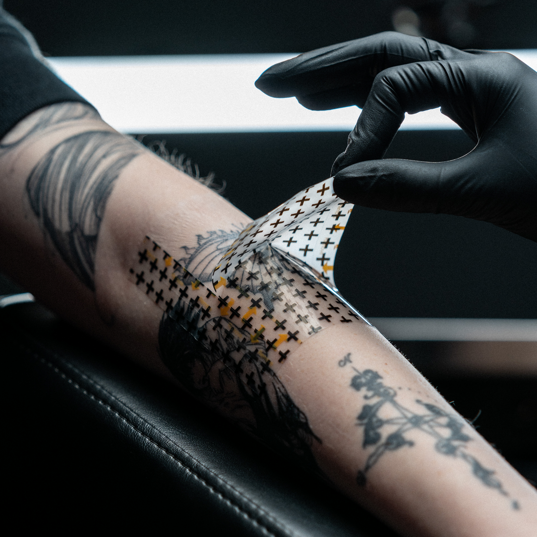 A tattoo artist wearing a black glove carefully peels a clear, cross-patterned aftercare bandage off a client's freshly tattooed forearm.