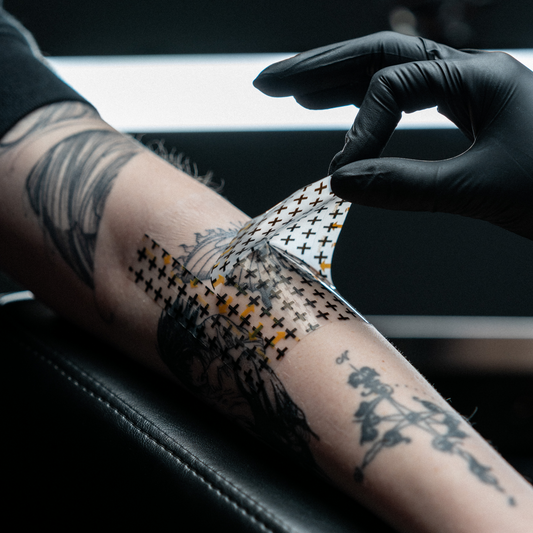 A tattoo artist wearing a black glove carefully peels a clear, cross-patterned aftercare bandage off a client's freshly tattooed forearm.