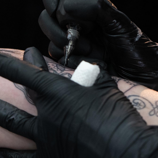 Close-up of an artist in black gloves executing detailed line work with a rotary tattoo machine onto a client's arm.